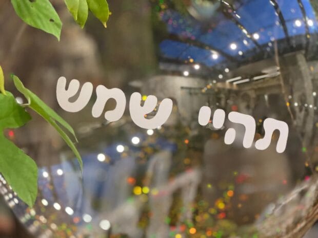 כיתוב בעברית תהיי שמש על חלון זכוכית בחלל עם צמחים ותאורה