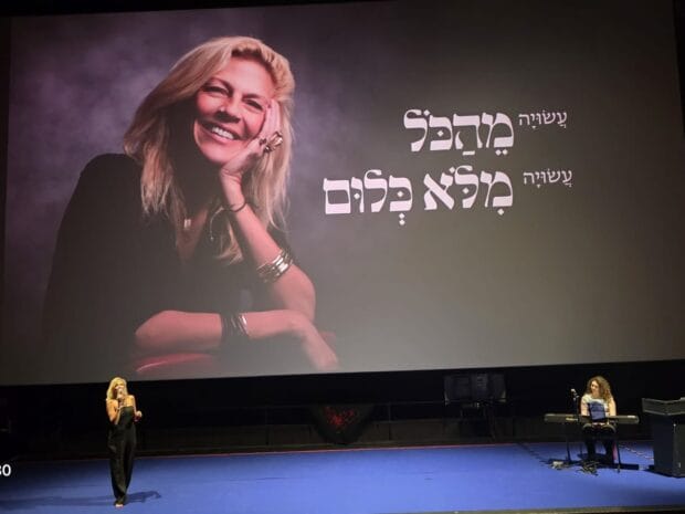אישה על במה ופסנתרנית מול מסך ענק עם דיוקן אישה מחייכת וכיתוב בעברית